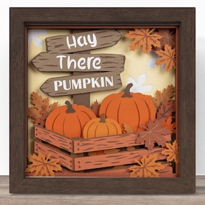 May include: A layered paper shadow box art piece in a dark brown frame featuring three orange pumpkins in a wooden crate, surrounded by autumn leaves and a signpost with the white text Hay There PUMPKIN against a light yellow background.