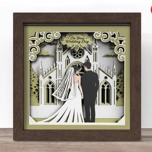 May include: A framed paper cut art piece featuring a bride and groom in front of a church. The artwork is layered with white and olive green paper, and the frame is brown. The text "On Your Wedding Day" is visible at the top.