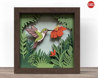 May include: A framed paper art piece featuring a hummingbird and a red flower. The hummingbird has green, white, and brown feathers. The artwork is set against a backdrop of green foliage and a light blue sky, all within a brown wooden frame.