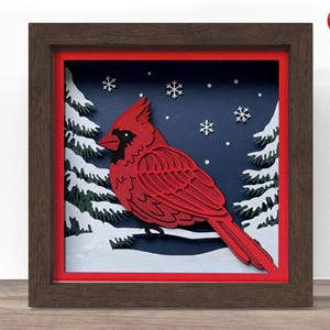 May include: A framed paper art piece depicting a red cardinal on a snowy branch, set against a dark blue backdrop with white snowflakes and evergreen trees. The frame is a dark brown colour, adding depth to the winter scene.