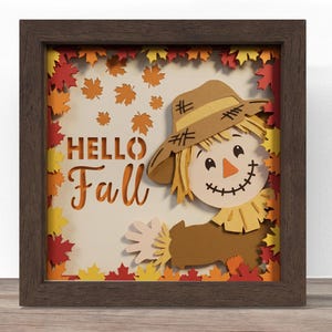 May include: A square shadow box frame with a dark wood finish containing a layered paper art piece. The design features a smiling scarecrow wearing a tan hat, surrounded by red, orange, and yellow maple leaves, with the text "HELLO Fall" in brown.