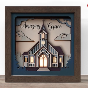 Puede incluir: Una obra de arte en papel enmarcada y multicapa con un diseño de iglesia. La obra de arte está en un marco marrón oscuro con un fondo azul. La iglesia es azul y blanca con las palabras "Amazing Grace" encima.