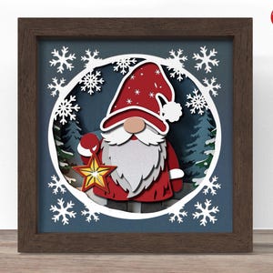 Puede incluir: Una obra de arte en papel enmarcada y en capas que presenta un gnomo navideño sosteniendo una estrella. El gnomo lleva un gorro y abrigo rojos con barba blanca. Copos de nieve blancos y una escena de bosque en el fondo.
