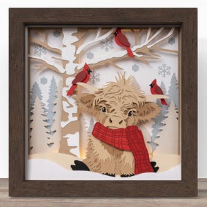 May include: A layered paper shadow box art piece in a dark brown frame featuring a tan Highland cow wearing a red plaid scarf. The background depicts a winter forest scene with white birch trees, evergreen trees, snowflakes, and three red cardinals.