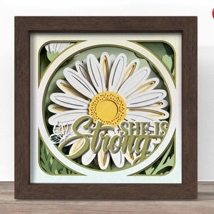 May include: A framed, layered paper art piece featuring a large daisy with white petals and a yellow center. The words "SHE IS Strong" are layered over the daisy. The frame is brown, and the background is green.