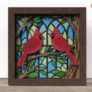 May include: A framed artwork featuring two red cardinal birds perched on a tree branch. The background has a stained-glass effect with blue, green, and white colours. The frame is a dark brown colour.