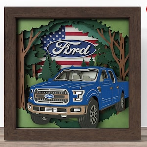 May include: A blue Ford pickup truck is framed in a wood frame with a papercraft scene behind it. The scene includes a forest, trees, and an American flag with the Ford logo on it.