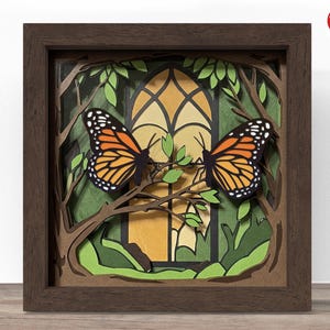 Puede incluir: Obra de arte en papel enmarcada y en capas, con dos mariposas monarca posadas en una rama. Las mariposas tienen alas naranjas y negras. El fondo incluye un diseño de vidriera y follaje verde. El marco es marrón oscuro.