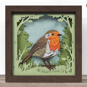 May include: A framed paper art piece depicting a robin. The bird has an orange breast, brown wings, and is set against a backdrop of green trees and foliage. The frame is a dark brown colour. The artwork is layered.