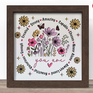May include: A framed floral art print with a brown wooden frame. The artwork features a central floral arrangement in pink, purple, and yellow, surrounded by text that reads "you are" and inspirational words like "strong" and "beautiful".