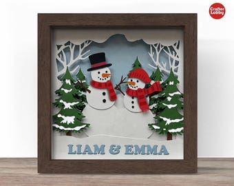 May include: A framed winter-themed shadow box featuring two snowmen. One snowman wears a black top hat and red scarf, the other a red knit cap and scarf. Green trees and white snow are layered in the frame. The text "LIAM & EMMA" is at the bottom.