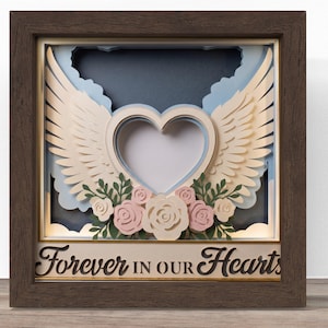 May include: A layered paper shadow box in a dark brown wooden frame featuring a central heart shape flanked by cream-colored angel wings. Below the heart are pink and cream paper roses with green leaves. The text Forever in our Hearts is displayed.