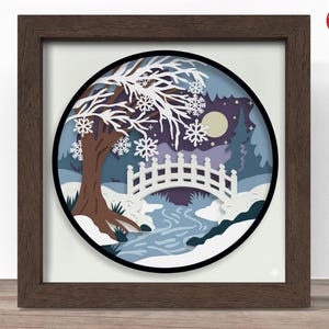 May include: A framed paper art piece depicting a winter scene. The artwork features a brown tree, a white bridge, and a river, all set against a backdrop of a snowy landscape and a full moon. The frame is a dark brown color.