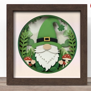 May include: A framed papercraft artwork featuring a green gnome with a white beard and a green hat. The gnome is surrounded by green foliage and two red and white mushrooms. The artwork is set in a brown wooden frame.