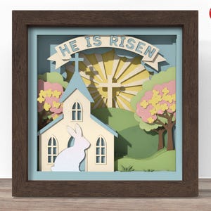 Puede incluir: Una obra de arte en papel enmarcada y en capas con un tema religioso. La obra presenta una iglesia, una cruz y las palabras "HE IS RISEN" en una pancarta. Un conejo blanco está frente a la iglesia. La paleta de colores incluye azul, amarillo, verde y marrón.