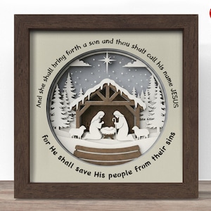 Könnte beinhalten: Ein gerahmtes Papierkunstwerk mit einer geschichteten Krippenszene. Das Kunstwerk zeigt einen Stall, Figuren und Schafe mit einem Stern darüber. Der Rahmen ist dunkelbraun, und der Text lautet: "Und sie wird einen Sohn gebären, und du sollst seinen Namen Jesus nennen, denn er wird sein Volk erlösen von ihren Sünden."