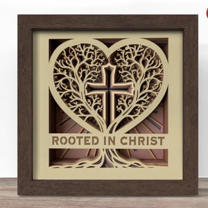 May include: A framed, layered wooden art piece featuring a heart-shaped tree design with a cross at the centre. The text "ROOTED IN CHRIST" is displayed at the bottom. The frame is brown.