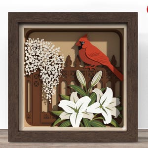 May include: A framed paper art piece featuring a red cardinal perched on a brown fence, with white lilies and a white floral element. The artwork is set against a beige background within a brown wooden frame.