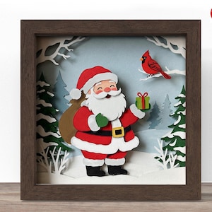 以下が含まれることがあります： 額装されたクリスマスのテーマのペーパーアート作品。赤いスーツと帽子を着たサンタクロースが、緑のプレゼントを持っています。赤いカージナルが枝に止まっています。背景には、木や枝のある雪景色が描かれています。フレームは茶色です。