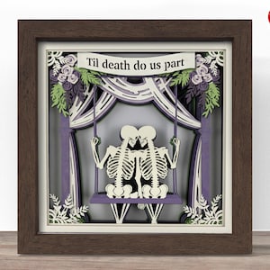 May include: A framed paper art piece with a layered design, depicting two skeleton figures on a swing. The artwork is in a wooden frame and includes the text "Til death do us part". The colour scheme features white, purple, and green.