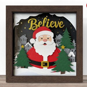 Puede incluir: Una pieza de arte en papel enmarcada con temática navideña, que presenta a Papá Noel con traje y gorro rojos, con la palabra "Believe" en letras doradas. La escena incluye árboles verdes, nieve y un pueblo, todo sobre un cielo nocturno oscuro.