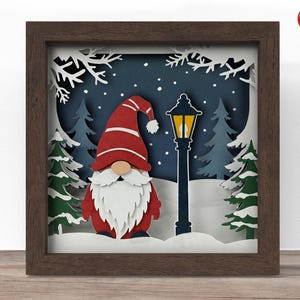 May include: A framed paper art piece depicting a gnome in a red hat and coat, with a white beard, set in a snowy winter scene with a lamppost and evergreen trees. The artwork is set in a dark brown wooden frame.
