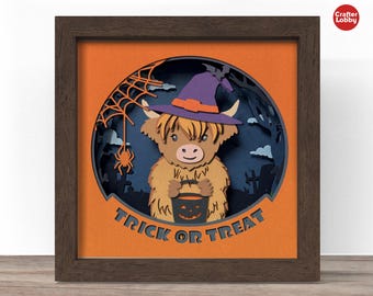 May include: A framed Halloween-themed paper art piece. It features a Highland cow wearing a witch hat and holding a jack-o'-lantern bucket. The words "Trick or Treat" are displayed at the bottom. The background includes a spider web and a graveyard scene.