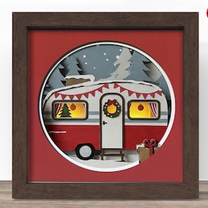 May include: A framed papercraft scene featuring a red and white camper decorated for Christmas. The camper has a wreath on the door and a garland with red pennants. The background shows a snowy landscape with pine trees.