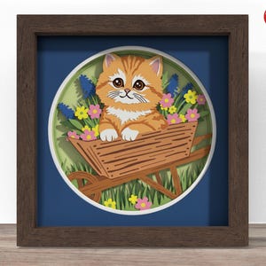 May include: A framed, layered paper art piece featuring an orange tabby kitten in a wooden wheelbarrow filled with flowers. The artwork is set against a blue background and framed in a dark brown wooden frame. The artwork is a charming, whimsical depiction of a cat in a garden.
