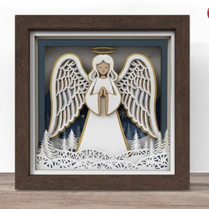 Può includere: Un'opera d'arte in carta a strati incorniciata con un angelo bianco con ali spiegate, un'aureola dorata e mani giunte. L'angelo è ambientato su uno sfondo invernale blu e bianco con alberi. La cornice è di colore marrone scuro.