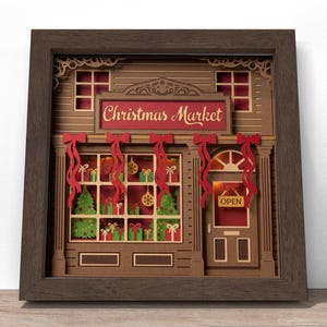 Puede incluir: Una obra de arte en papel en capas enmarcada que representa una fachada de mercado navideño. El letrero sobre la tienda dice "Christmas Market" en rojo. Las ventanas muestran árboles de Navidad, regalos y adornos. Lazos y cintas rojas decoran la fachada.