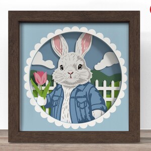 May include: A framed paper art piece featuring a white rabbit wearing a blue denim jacket and holding a pink tulip. The rabbit is set against a blue backdrop with a white picket fence and clouds. The frame is brown.