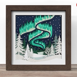 Op de afbeelding: Een ingelijst papieren kunstwerk dat het noorderlicht uitbeeldt. Het kunstwerk toont lagen papier gesneden in een winterbosscène met groene en witte bomen, een besneeuwde grond en een donkerblauwe lucht met groene auroralichten. De lijst is donkerbruin.