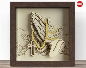 May include: A 3D papercraft of a pair of hands clasped in prayer with a rosary and a cross. The hands are made of beige paper and the rosary is made of gold paper. The cross is made of brown paper. The background is a light beige color.