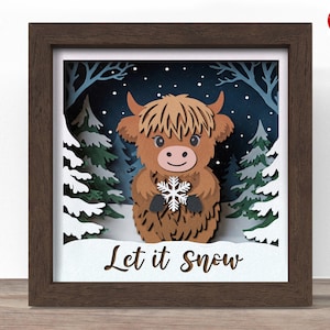 May include: A framed paper art piece depicting a brown Highland cow holding a snowflake in a snowy forest setting. The artwork includes the text "Let it Snow" and is encased in a dark brown wooden frame.