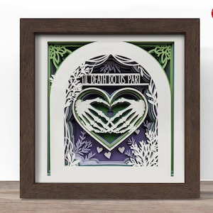 May include: A framed, layered paper art piece with a dark brown wooden frame. The artwork features a heart formed by skeleton hands, with the words "Till Death Do Us Part" above. The background includes green, white, and purple layers.