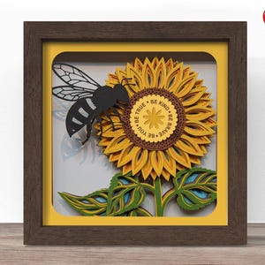 May include: A layered paper art piece featuring a sunflower and a bee. The sunflower is yellow and brown with green leaves and stem. A black bee is positioned on the flower. The artwork is framed in a brown wooden shadow box with the words "BE TRUE, BE KIND, BE BRAVE, BE YOU".