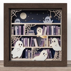 Puede incluir: Un marco de caja de sombra cuadrado que contiene una escena de manualidades en papel multicapa de una estantería llena de libros morados y blancos. Varias figuras de fantasmas blancos se sitúan entre los estantes, con detalles de telarañas.