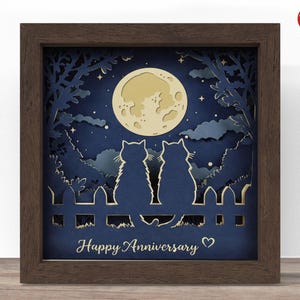 May include: A framed paper art piece depicting two cat silhouettes on a fence, beneath a full moon and starry night sky. The artwork is layered with a dark blue background and a brown wooden frame. The text "Happy Anniversary" is at the bottom.