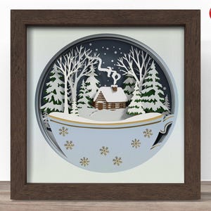 Könnte beinhalten: Gerahmtes Papierkunstwerk, das eine Winterlandschaft in einer Teetasse darstellt. Die Szene zeigt eine schneebedeckte Hütte, Bäume und Schneeflocken. Die Teetasse ist hellblau mit goldenen Akzenten, und der Rahmen ist dunkelbraun.