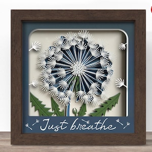 May include: A framed paper art piece featuring a layered dandelion design in shades of blue and white. The artwork includes green leaves and the phrase "Just breathe" in white script. The frame is a dark brown color.