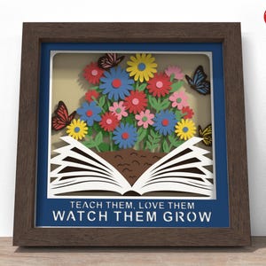 May include: A framed paper art piece featuring a bouquet of colorful paper flowers and butterflies. The flowers are blue, yellow, pink, and red. The open book at the bottom has the words "TEACH THEM, LOVE THEM, WATCH THEM GROW".