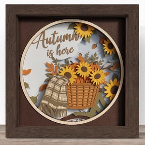 May include: A square dark brown wooden shadow box frame containing a layered paper art piece. The circular design features a basket of sunflowers and pumpkins, a plaid blanket, and the text "Autumn is here" in a script font against a white background.