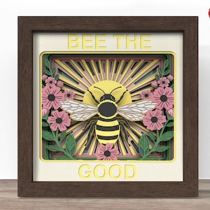 May include: A framed, layered paper art piece with a bee design. The artwork features a yellow and black bee in front of a sunburst design, surrounded by pink flowers and the words "BEE THE GOOD". The frame is brown.