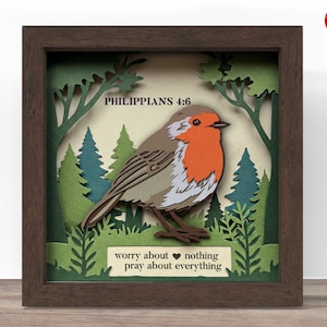 Puede incluir: Una obra de arte en papel enmarcada con un diseño en capas de un pájaro, árboles y el texto "Filipenses 4:6" y "no se inquieten por nada; más bien, en toda ocasión, con oración y ruego, presenten sus peticiones a Dios." El pájaro tiene pecho rojo y alas marrones. El marco es marrón.