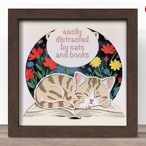 May include: A framed paper art piece depicting a sleeping tabby cat on a book, with flowers and the text "easily distracted by cats and books". The artwork is set against a dark blue backdrop, with a brown frame. The measurements are in centimeters.