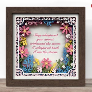 May include: A framed paper art piece with a dark brown wooden frame. The artwork features layered paper cutouts of flowers in pink, yellow, blue, and white, surrounding the text "They whispered you cannot withstand the storm I whispered back I am the storm."
