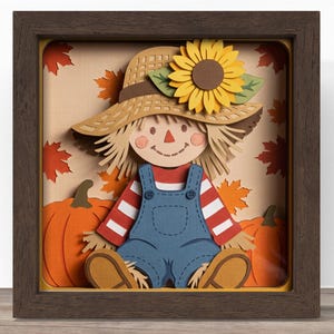 May include: A layered paper shadow box art piece featuring a scarecrow wearing a straw hat with a yellow sunflower, a red and white striped shirt, and blue overalls. The background displays orange pumpkins and scattered red maple leaves.