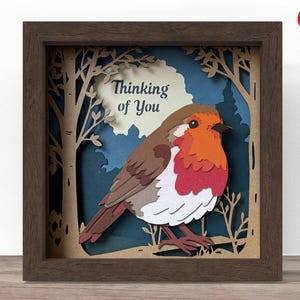 May include: A framed paper art piece featuring a colorful robin bird with the text "Thinking of You." The bird has a red breast, orange head, and brown wings, set against a layered forest scene. The frame is a dark brown colour.