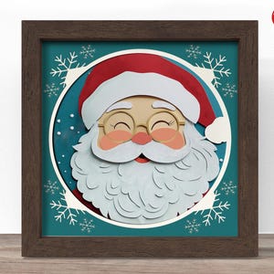 Puede incluir: Una obra de arte en papel enmarcada y en capas que presenta a Papá Noel. Papá Noel tiene un gorro rojo, mejillas rosadas y una barba blanca. El fondo es turquesa con copos de nieve blancos y un círculo blanco alrededor de la cara de Papá Noel. El marco es marrón.